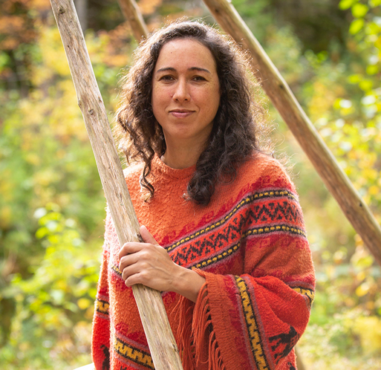 A woman with shoulder-length curly hair is standing in a partially-build wigwam against a backdrop of green leaves