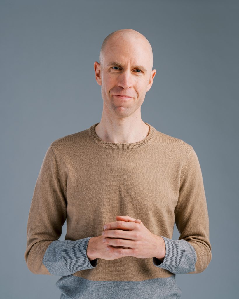 Headshot of Joel Berbaum, a bald white man wearing a beige long-sleeved shirt with blue cuffs.